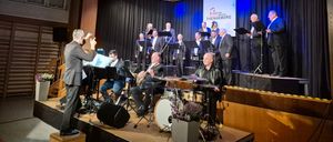 Eine Gruppe von Musikern tritt auf einer Bühne auf, mit einem Banner dahinter. Die Bühne hat Instrumente und Lautsprecher. Das Publikum ist nicht sichtbar.