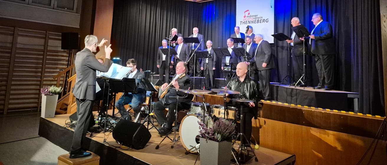 Eine Gruppe von Musikern tritt auf einer Bühne auf, mit einem Banner dahinter. Die Bühne hat Instrumente und Lautsprecher. Das Publikum ist nicht sichtbar.