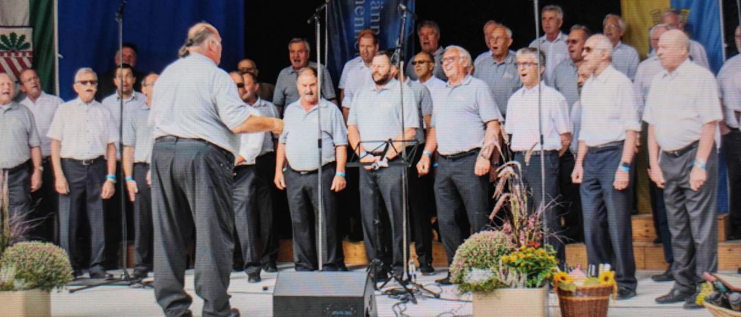 Ein Chor von Männern in hellblauen Hemden und dunklen Hosen tritt auf der Bühne auf, hinter Mikrofonen stehend. Ein älterer Mann führt sie an, während andere singen. Im Hintergrund befindet sich ein blauer Banner mit weißem Text.