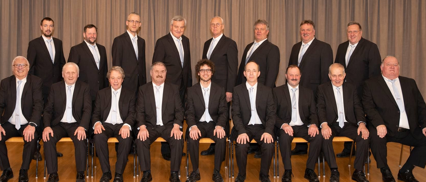 Eine Gruppe formell gekleideter Männer, die in einer Reihe sitzen, möglicherweise für ein Foto posieren, mit einem Vorhang dahinter.