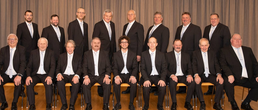 Eine Gruppe formell gekleideter Männer, die in einer Reihe sitzen, möglicherweise für ein Foto posieren, mit einem Vorhang dahinter.