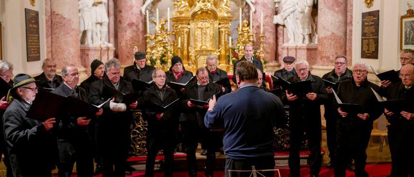A choir dressed in black is singing in a church. A man stands before them with a music stand. The choir sits in front of a golden altar adorned with candles and statues.