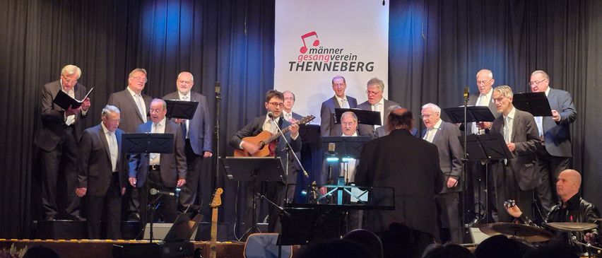 Ein Chor tritt auf der Bühne auf, mit einem Banner hinter ihnen, das 'Mannen Gesangverein Thennberg' besagt. Die Chormitglieder tragen Anzüge und Krawatten, und einer spielt Gitarre. Vor ihnen stehen Notenständer und Instrumente. Das Publikum beobachtet sie.