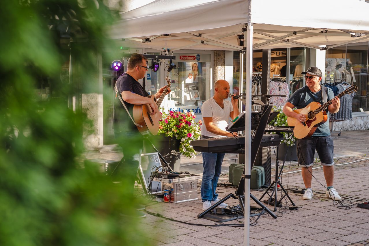 Drei Männer spielen auf einer Bühne mit einem weißen Baldachin. Ein Mann spielt Gitarre, ein anderer spielt Keyboard und der dritte singt in ein Mikrofon. Ein Topf steht vor der Bühne.