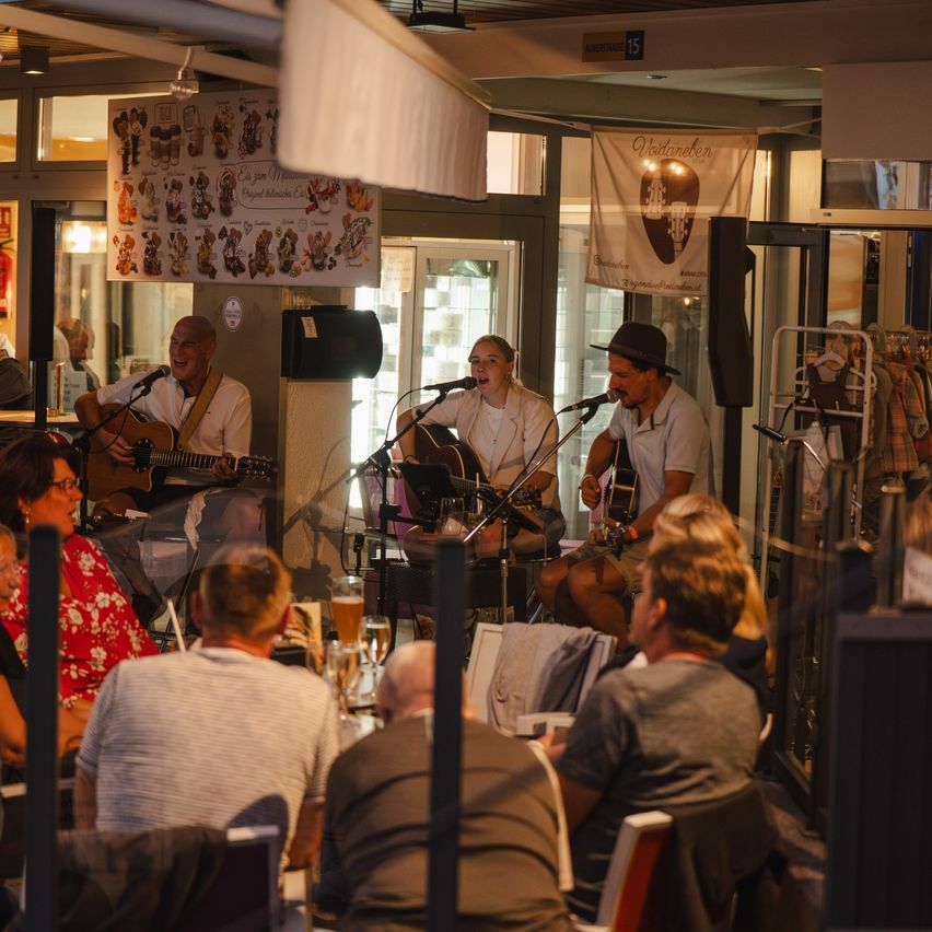 Ein Trio von Musikern tritt in einem Restaurant mit Publikum auf. Sie spielen Gitarren und singen. Eine Fahne hängt an der Wand.