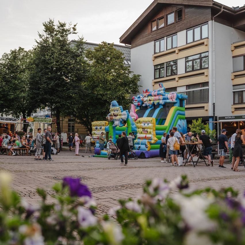 Menschen versammeln sich um ein buntes aufblasbares Hüpfburg in einem Stadtplatz, mit Bäumen und Gebäuden im Hintergrund.