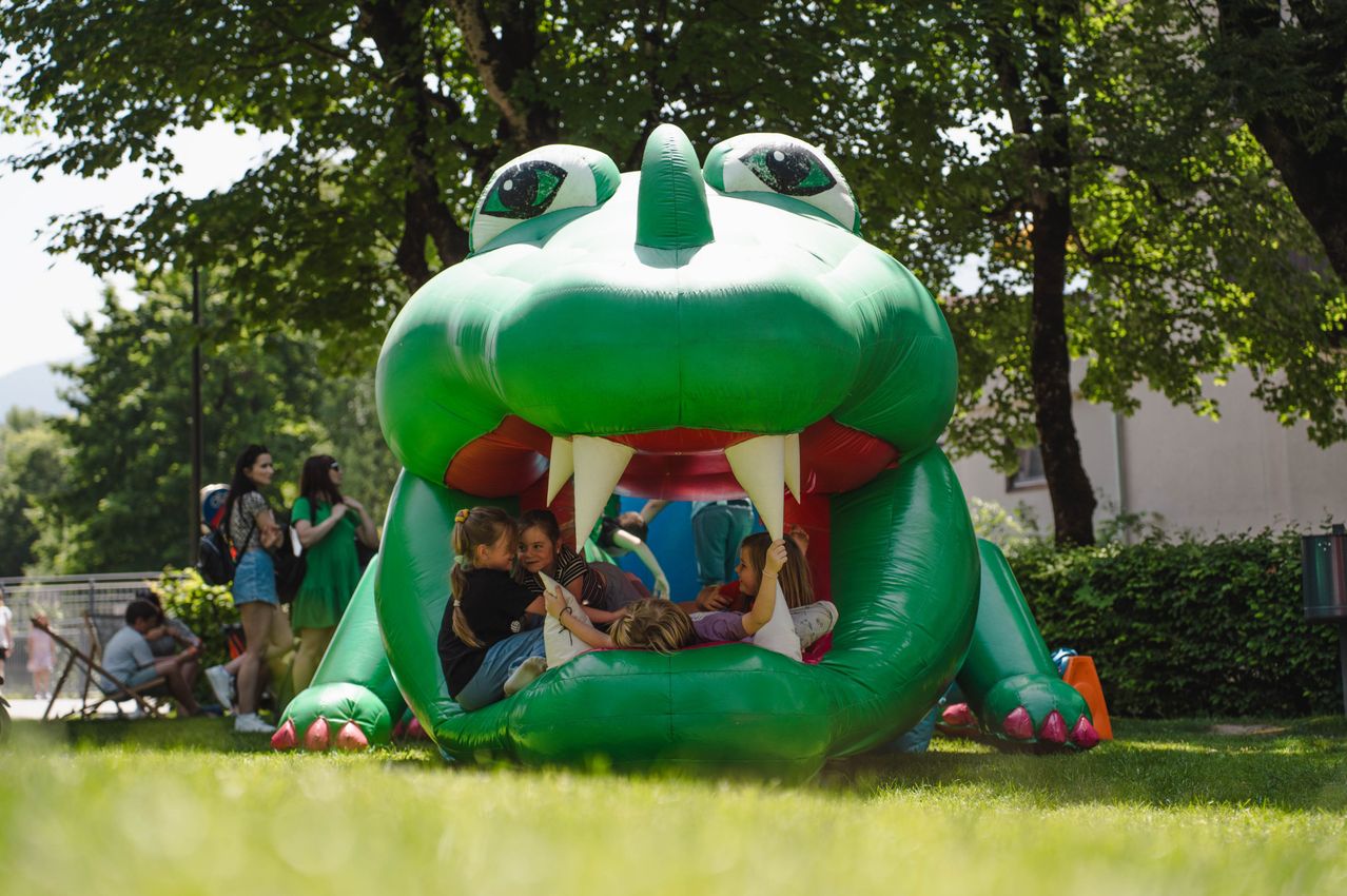 Vier Kinder spielen in einem grünen aufblasbaren Krokodil im Freien. Sie liegen auf dem Bauch und schauen aus dem Maul des Krokodils. Bäume und ein Gebäude sind im Hintergrund.