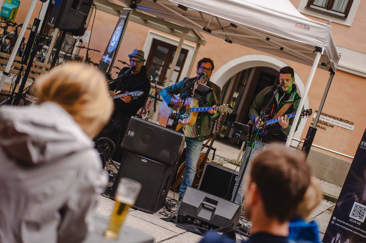 Drei Männer spielen Gitarre und singen auf einer Bühne unter einem weißen Zelt. Sie sind von Lautsprechern umgeben und ein Publikum schaut zu.