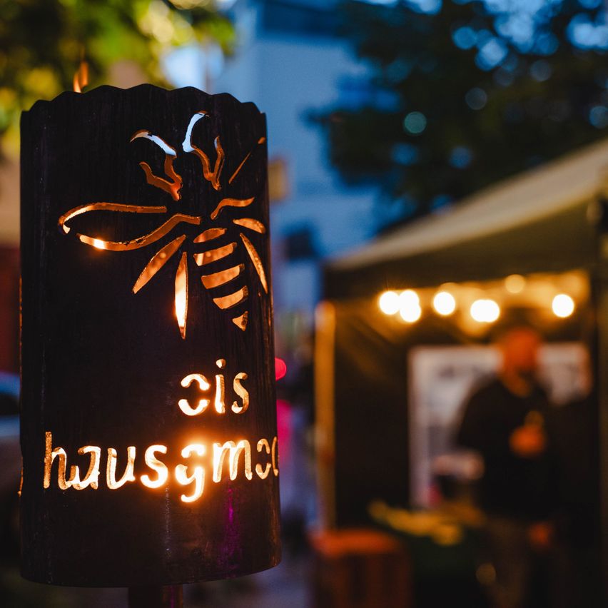 Ein hölzerner Pfosten mit einem Bienendesign und den Worten 'zis hausgmai' ist beleuchtet. Im Hintergrund gibt es ein verschwommenes Bild von einer Person und einem Tisch mit Lichterketten.