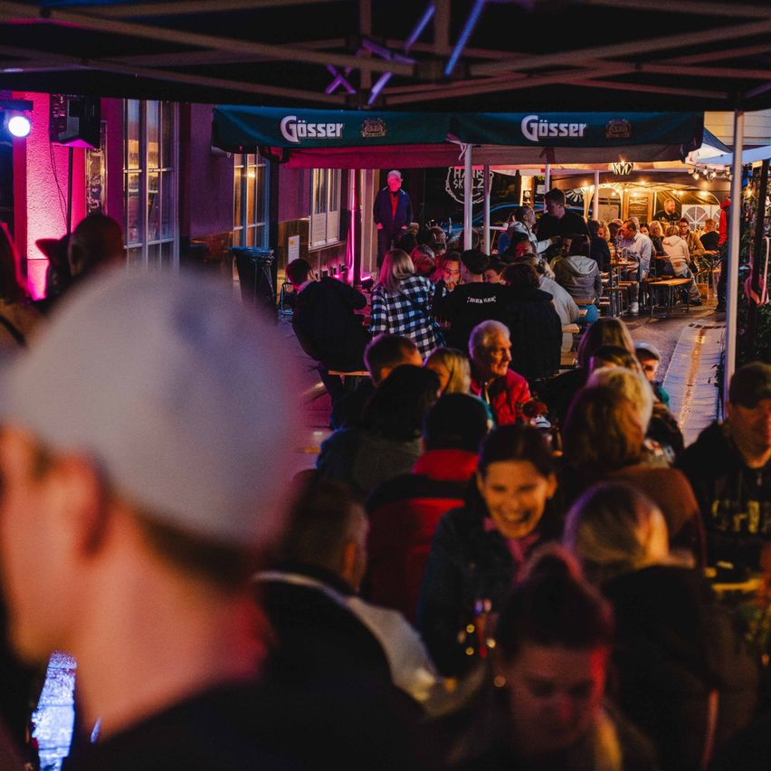 Ein geschäftiges Outdoor-Event bei Nacht, mit Menschen unter Gosser-Zelten versammelt, ein Mann spricht in ein Mikrofon und andere sitzen an Tischen.