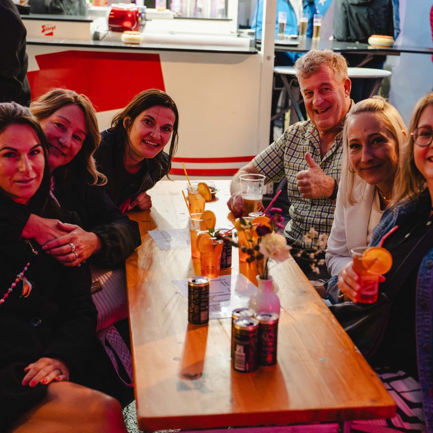 Eine Gruppe von Menschen, darunter Frauen und Männer, sitzen um einen Tisch in einer Bar. Sie lächeln und halten Getränke. Hinter ihnen steht eine Theke mit Gegenständen.