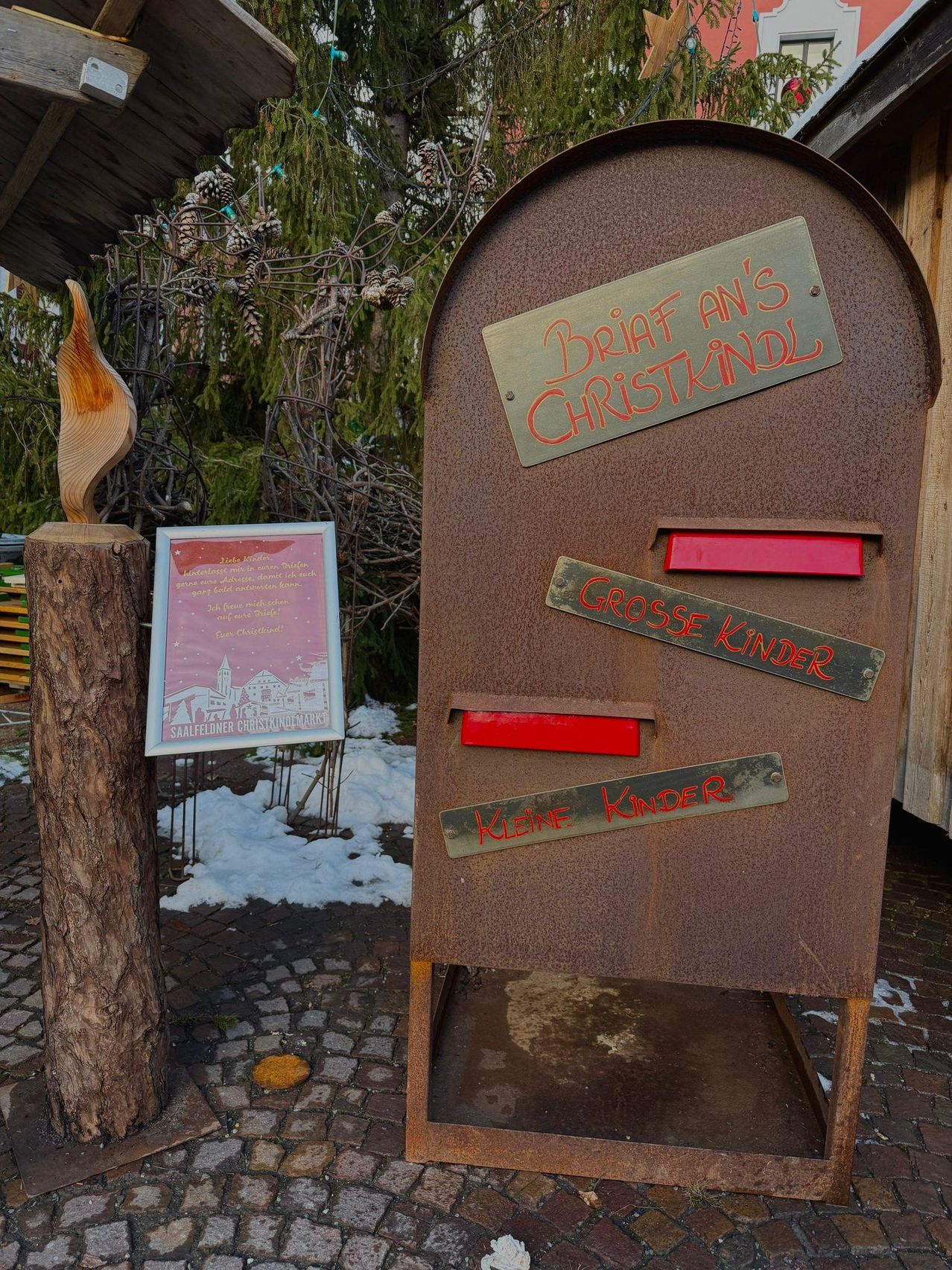 Ein Briefkasten für Weihnachtsbriefe an das Christkind mit separaten Bereichen für große und kleine Kinder steht in einem verschneiten Bereich. Ein Schild sagt 'Brief für Christkindl' und ein anderes 'Grosse Kinder' und 'Kleine Kinder'.