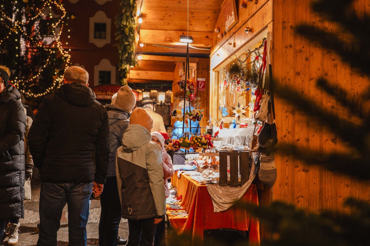 Eine Gruppe von Menschen erkundet einen festlichen Markt mit Holzständen und Dekorationen. Ein Tisch zeigt verschiedene Ornamente.