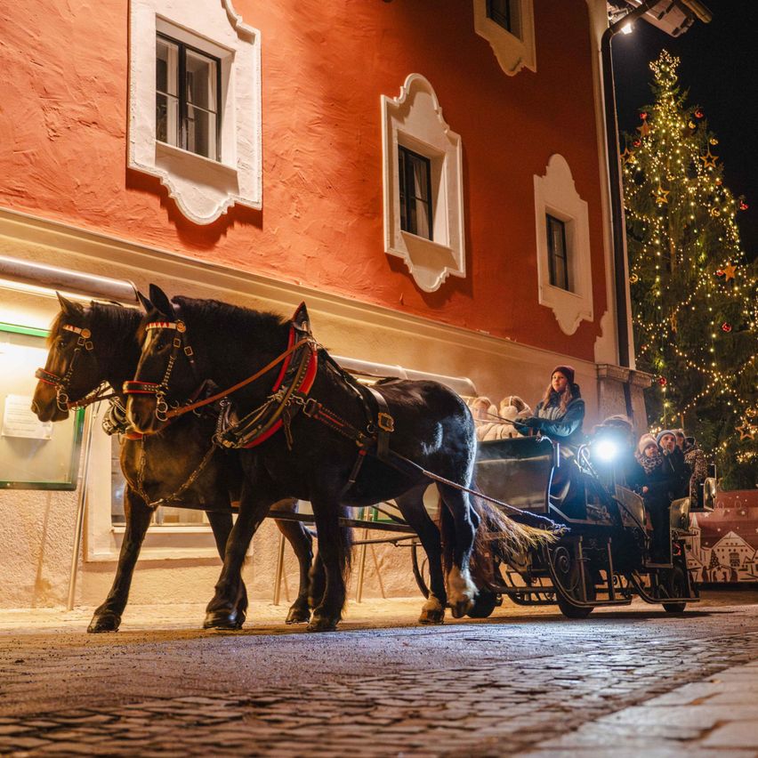 Eine Pferdekutsche mit einer Frau und Kindern fährt nachts durch eine festlich geschmückte Straße. Die Kutsche wird von einem Licht beleuchtet.