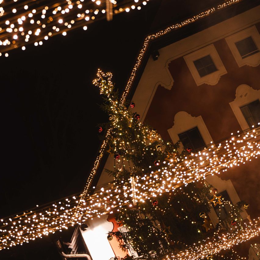 Ein Weihnachtsbaum ist mit Lichterketten beleuchtet und hängt über einem Gebäude. Der Baum ist mit roten Ornamenten geschmückt. Das Gebäude hat eine Holzfassade und mehrere Fenster.