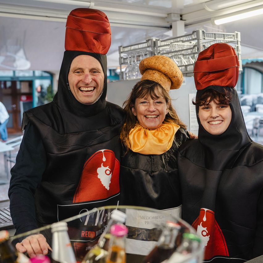 Drei Menschen in roten Hüten und schwarzen Kostümen posieren für ein Foto, lächeln in einem Lebensmittelstand mit Flaschen und Essensartikeln vor ihnen.