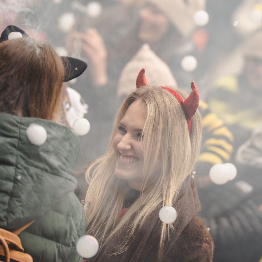Eine Frau mit Teufelshörnern lächelt, während sie mit einer anderen Person interagiert, die eine Katzenohren-Haarspange trägt. Sie sind von anderen umgeben, möglicherweise bei einer Halloween-Veranstaltung.