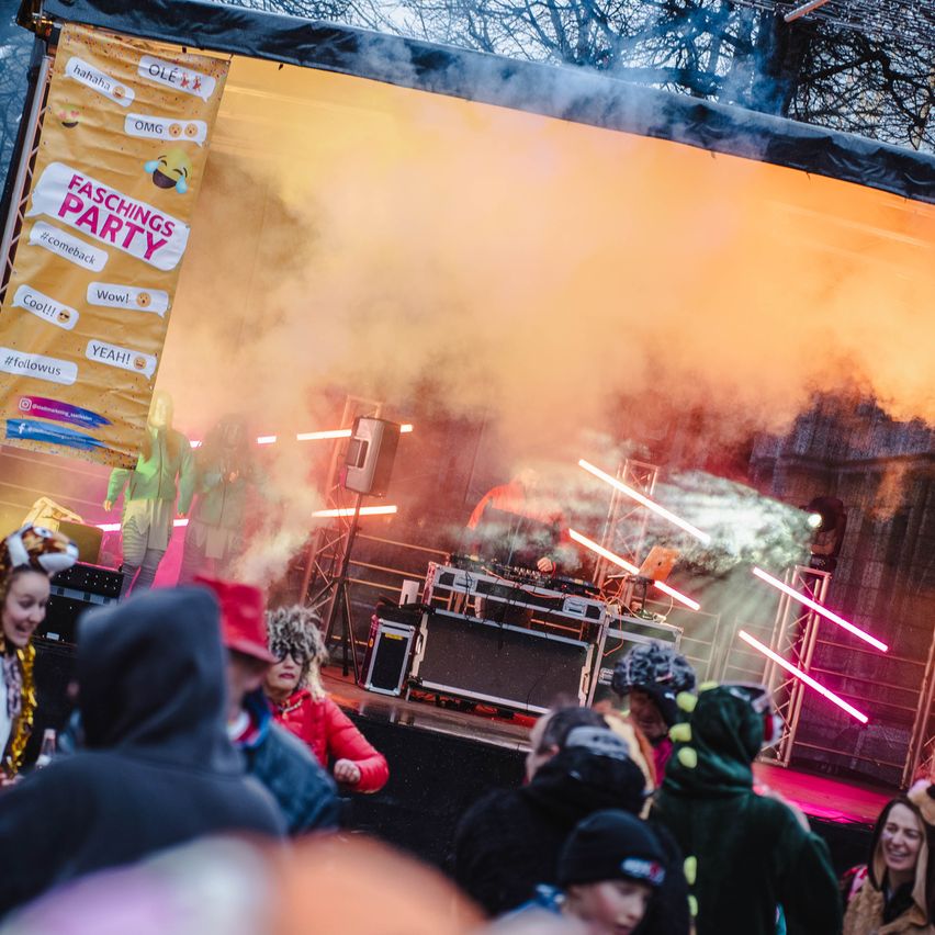 Eine lebhafte Musikveranstaltung mit Raucheffekten auf der Bühne, einem Banner mit der Aufschrift 'Faschings Party' und einer Menge Menschen, die die Show genießen.