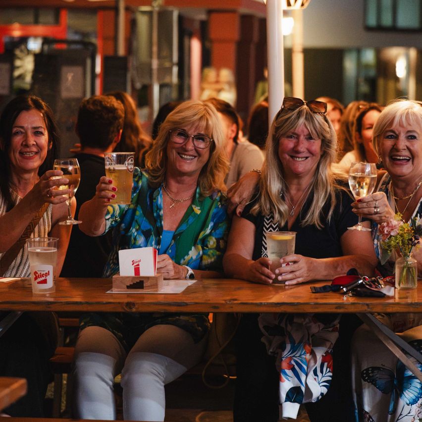 Vier Frauen sitzen an einem Tisch, lächeln und halten Gläser mit Bier und Wein. Sie befinden sich in einer lebhaften sozialen Umgebung mit anderen Menschen im Hintergrund.