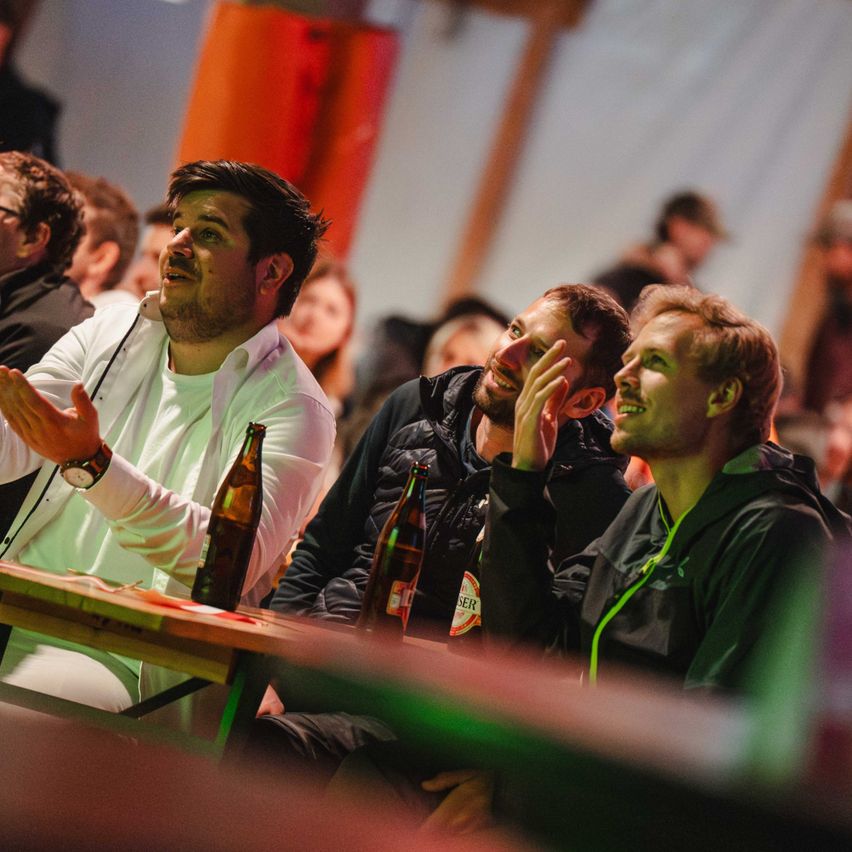 Vier Männer sitzen an einem Tisch mit Bierflaschen. Der Mann in Weiß klatscht. Der Mann in Schwarz lächelt und berührt sein Kinn.