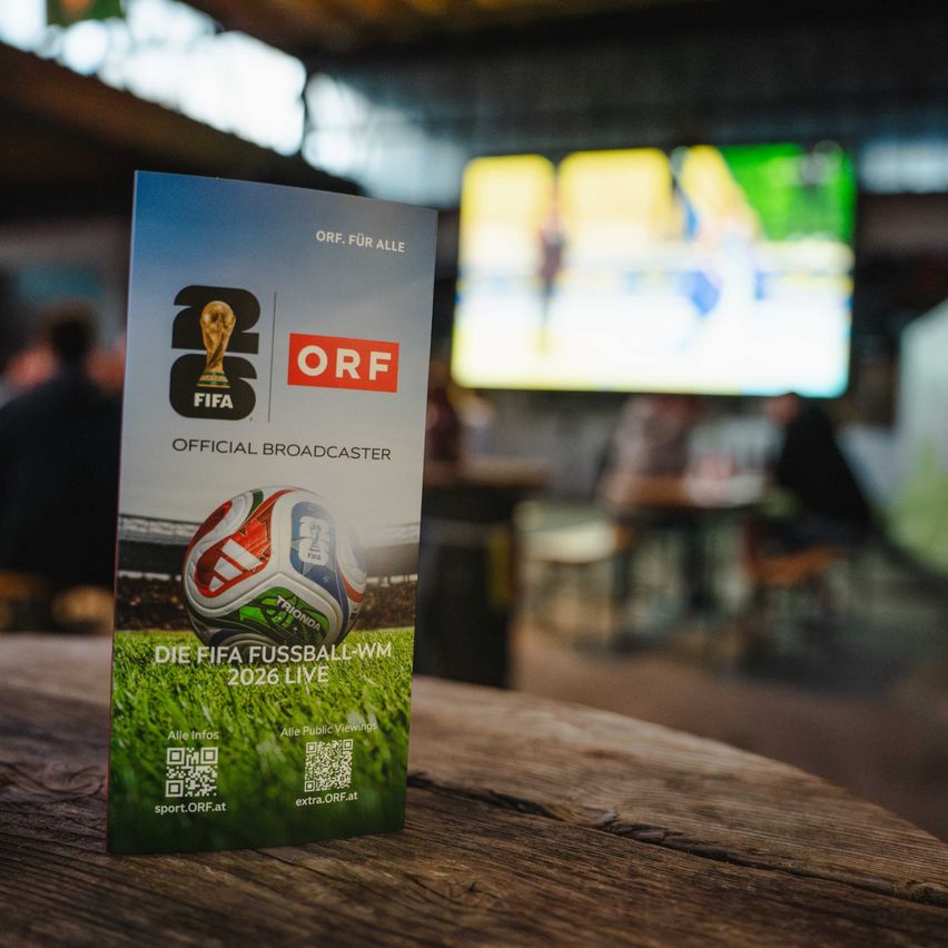 Plakat für die Übertragung der FIFA-WM 2026 auf ORF. Es zeigt einen Fußball und einen großen Bildschirm mit einem Fußballspiel im Hintergrund.