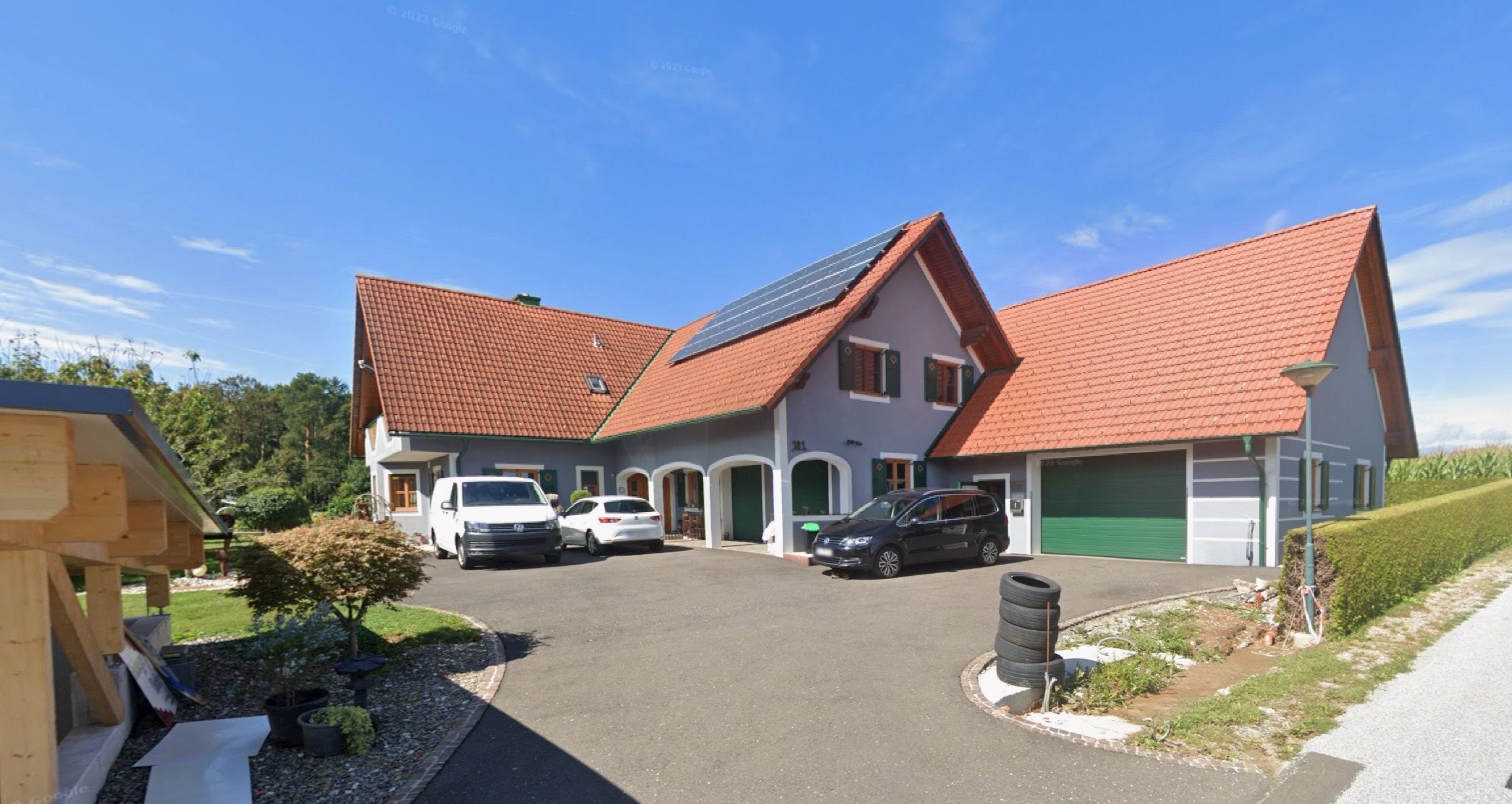 Ein modernes Haus mit Solarpanelen auf dem Dach und drei Autos auf der Einfahrt. Das Haus hat ein rotes Dach, grüne Türen und eine grüne Garagentür.