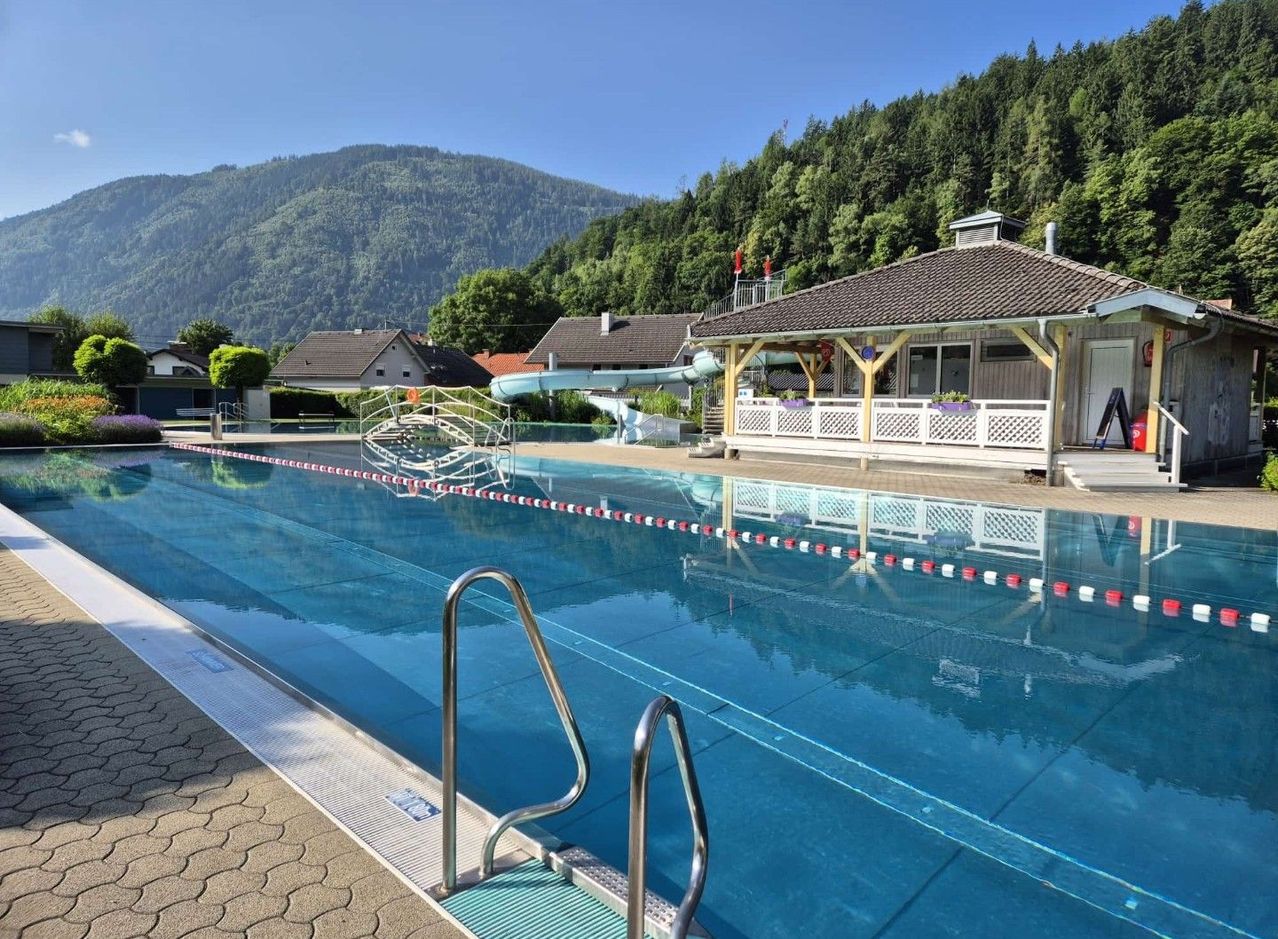Ein Freibad mit rot-weißer Bahnlinie, einer Rutsche und einem kleinen Gebäude mit Veranda. Im Hintergrund befinden sich Berge und Bäume.