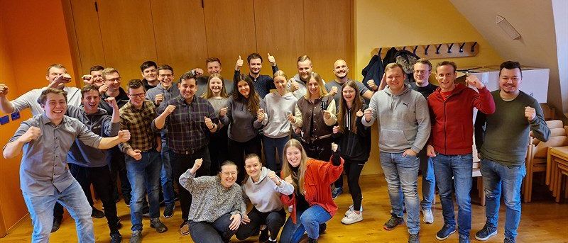 Eine Gruppe von Menschen posiert für ein Foto in einem Raum, einige sitzen, andere stehen, alle lächeln und zeigen den Friedenszeichen.