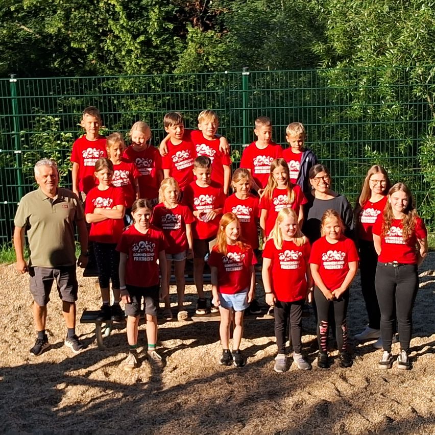 Eine Gruppe von Kindern und Erwachsenen in roten Shirts posiert für ein Foto im Freien. Dahinter steht ein grüner Zaun mit Bäumen.
