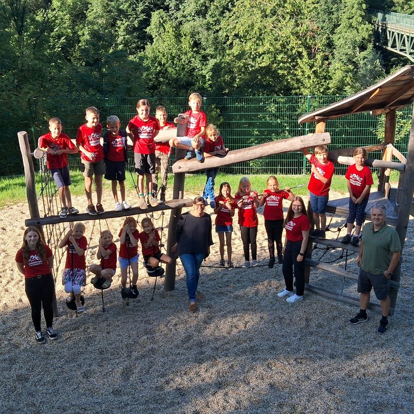 Eine Gruppe junger Menschen in roten T-Shirts steht und spielt auf einer Holzkonstruktion in einem Park. Einige sitzen auf Schaukeln, während andere auf einer Wippe stehen. Eine Frau und ein Mann stehen davor. Bäume sind im Hintergrund.