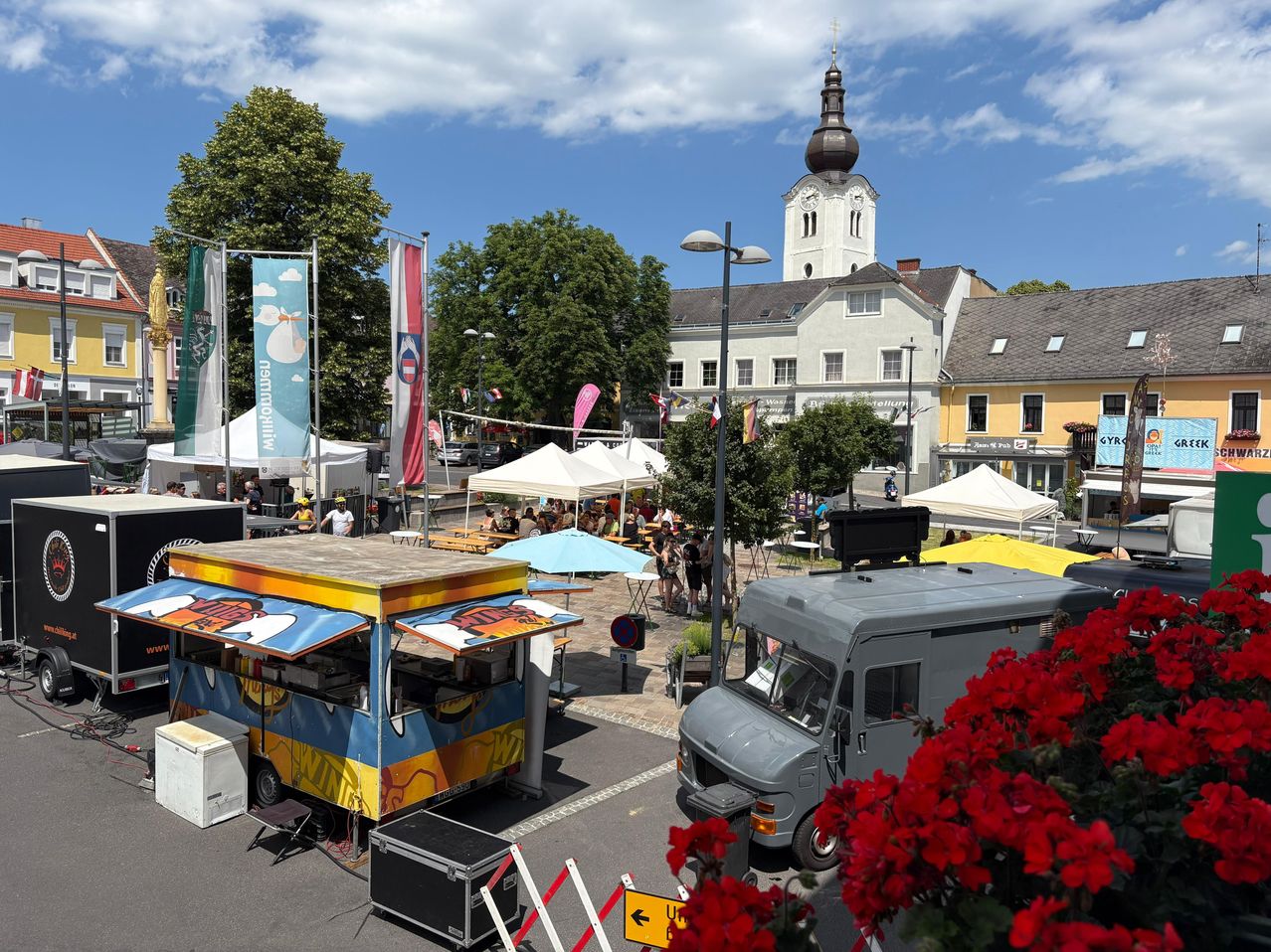 Ein Food-Truck-Festival mit bunten Food-Trucks und Zelten unter blauem Himmel, mit einer Kirche und Gebäuden im Hintergrund.