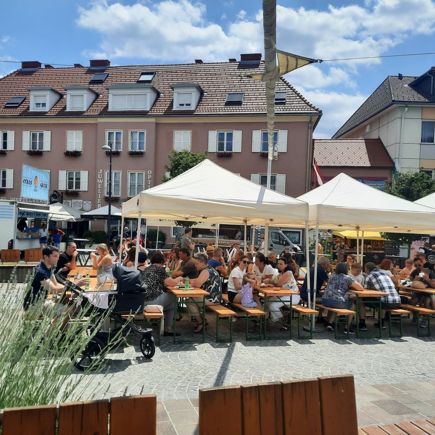 Menschen sitzen unter weißen Zelten auf einer Kopfsteinpflasterstraße an Tischen. Dahinter stehen Gebäude, eines mit einer Flagge und ein anderes mit einem Schild, das Juwelier liest. Bäume und ein bewölkter Himmel sind im Hintergrund zu sehen.