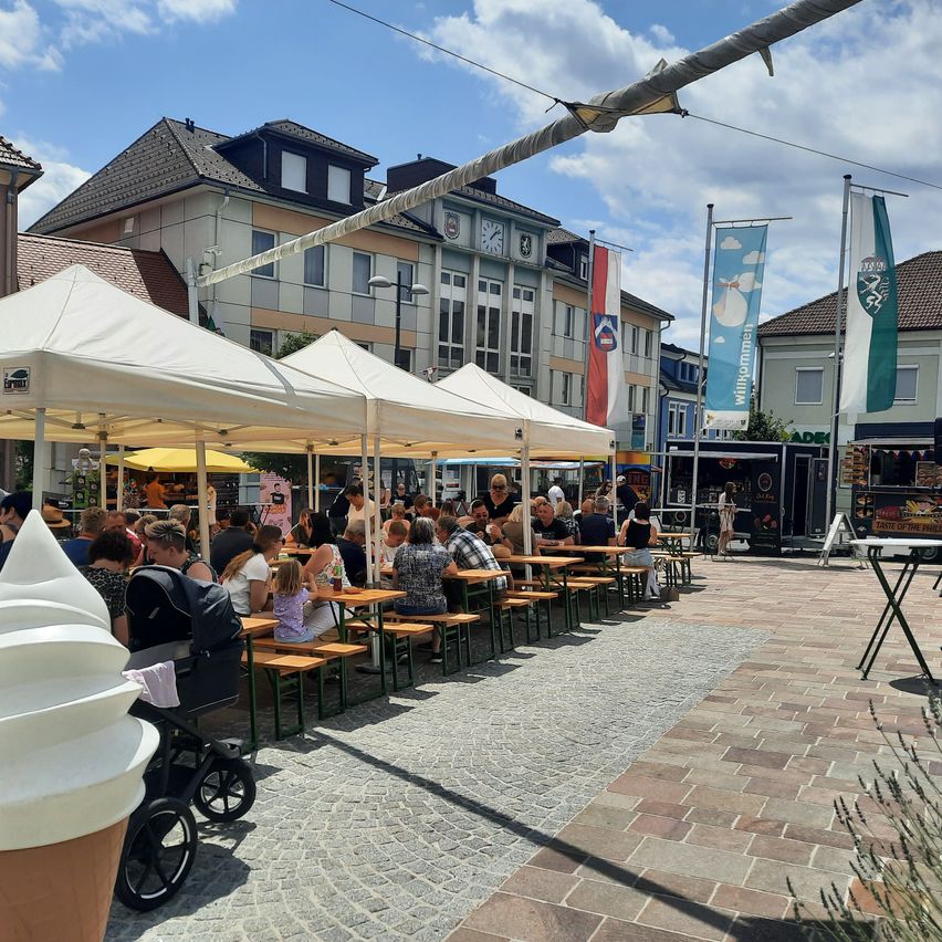 Ein Outdoor-Essbereich mit weißen Zelten und Tischen. Mehrere Menschen sitzen, einige im Schatten. Im Hintergrund sind Gebäude und Straßenlaternen zu sehen.