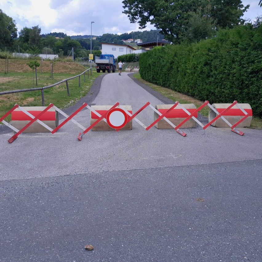 Eine Straße ist mit roten und weißen Barrieren gesperrt, mit einer Person, die geht, und einem Lkw im Hintergrund, umgeben von Grün und einem Zaun.