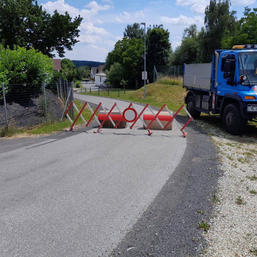 Ein blauer Lkw ist am Straßenrand geparkt, vor ihm stehen Barrieren. Die Straße ist von Gras und Bäumen umgeben. Ein Straßenlicht steht in der Nähe.