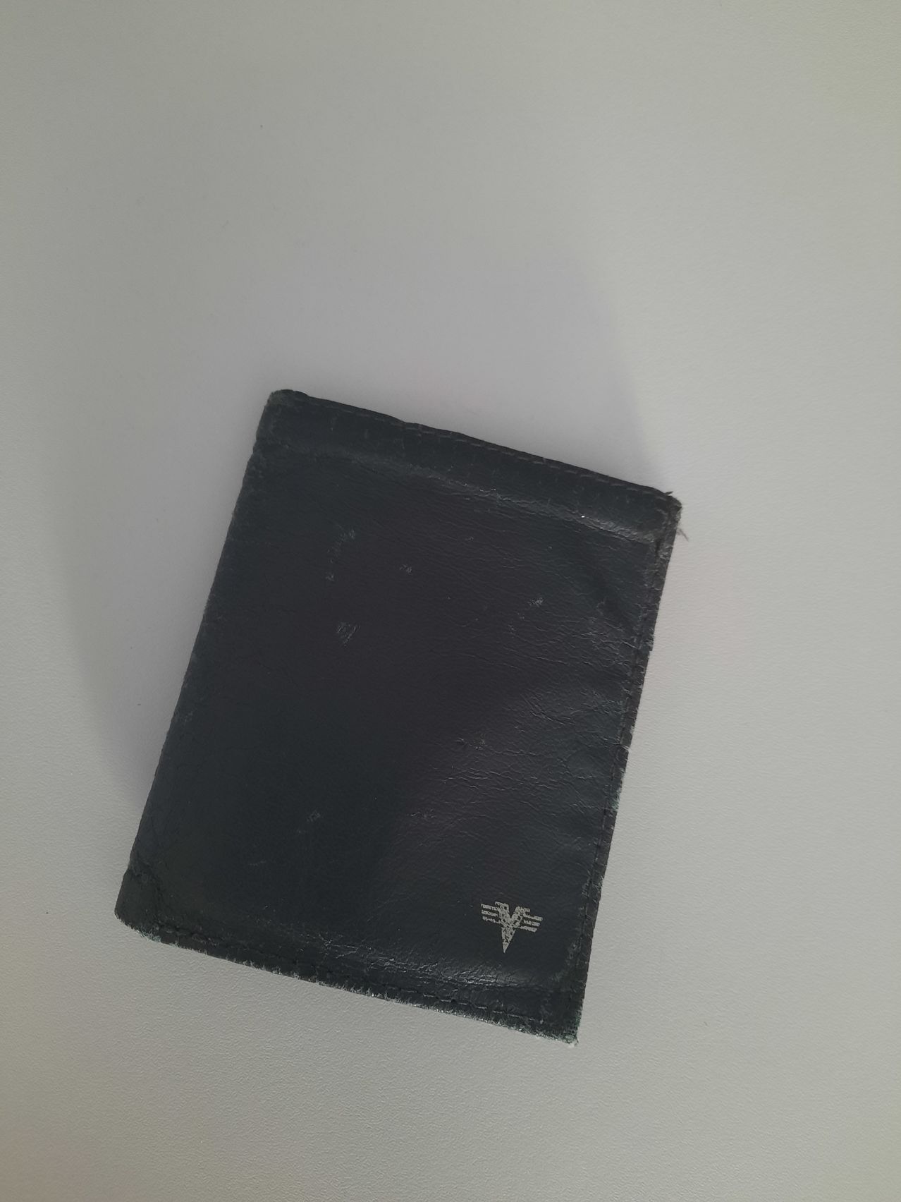 Ein schwarzer Lederbrieftasche mit einem silbernen Logo auf der Vorderseite, mit Anzeichen von Abnutzung, liegt auf einer weißen Oberfläche.