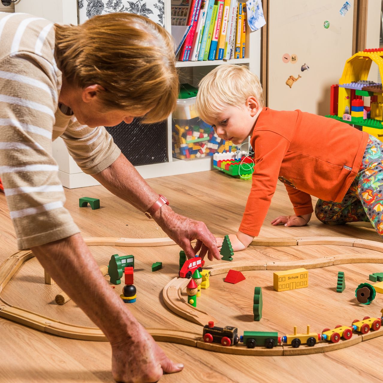 Eine ältere Frau und ein junger Junge spielen mit einem Holzeisenbahn-Set auf einem Holzboden.