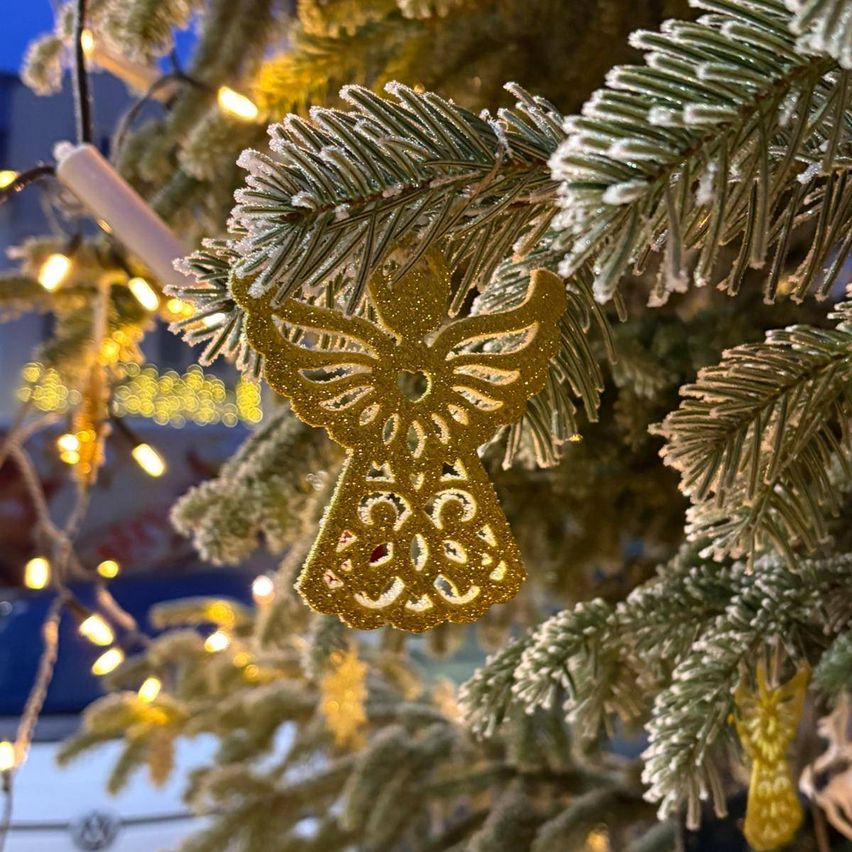 Ein goldener Engel-Schmuck hängt an einem vereisten Weihnachtsbaum mit leuchtenden Lichtern im Hintergrund.