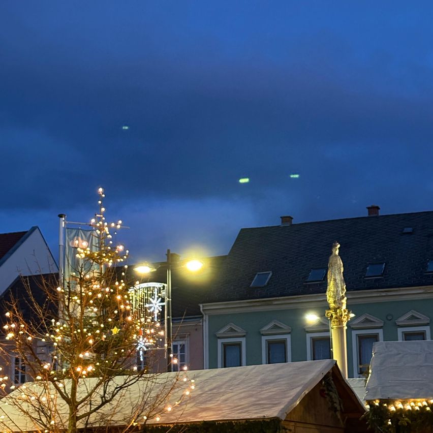 Ein beleuchteter Weihnachtsbaum steht vor einem Gebäude mit einer Statue, unter einem blauen Himmel mit Wolken.