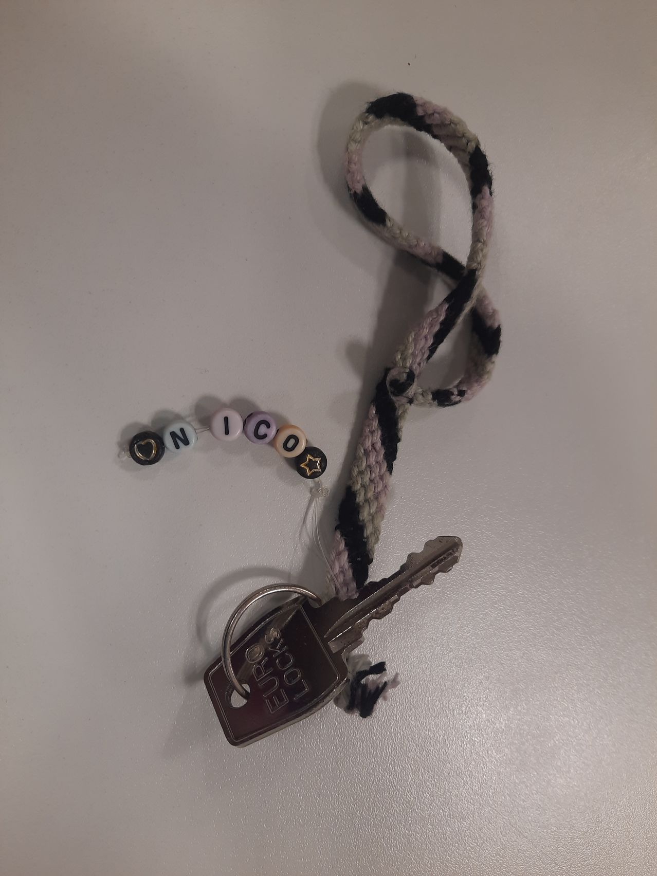 Ein Schlüssel mit einem Euro-Schloss-Tag ist an einer rosa-schwarzen gewebten Schlüsselkette befestigt. Die Schlüsselkette hat Buchstaben, die 'ON ICO' buchstabieren, mit einigen Perlen.
