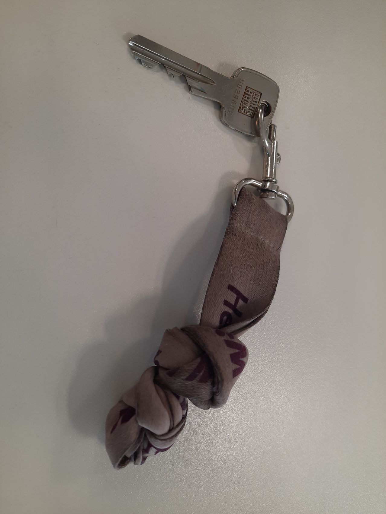 Ein Schlüsselanhänger mit einem grauen Schlüsselband und den Buchstaben 'HEW' darauf, an einem silbernen Schlüssel befestigt.