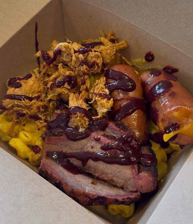 Eine Schachtel gefüllt mit geräuchertem Fleisch, ein paar Scheiben Brisket und etwas geriebenes Fleisch mit Soße obenauf. Es gibt auch zwei Würste, Mac and Cheese und einige gelbe Käsescheiben.
