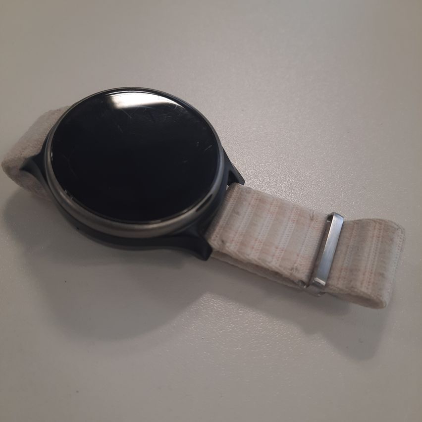 A black smartwatch with a black face and a beige strap with a silver buckle is placed on a white surface.