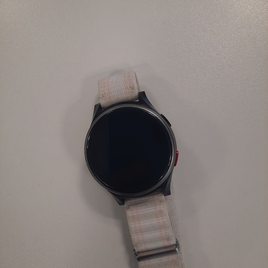 A black smartwatch with a round face and a red button is placed on a white surface. The watch has a white strap with a checkered pattern.