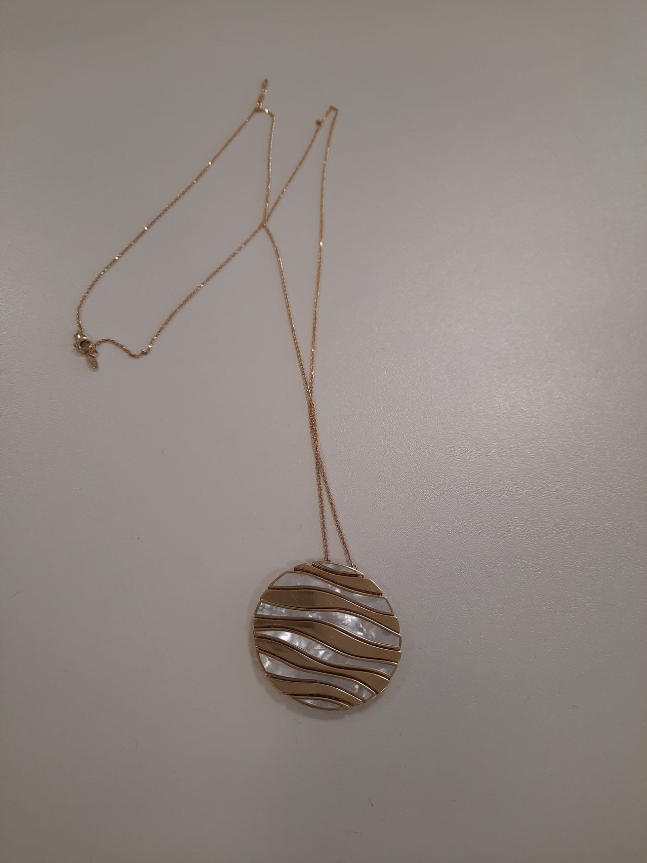 Eine Goldkette mit einem runden Anhänger, der mit wellenförmigen Mustern aus Perle und Gold verziert ist. Der Anhänger hängt an einer langen Goldkette.