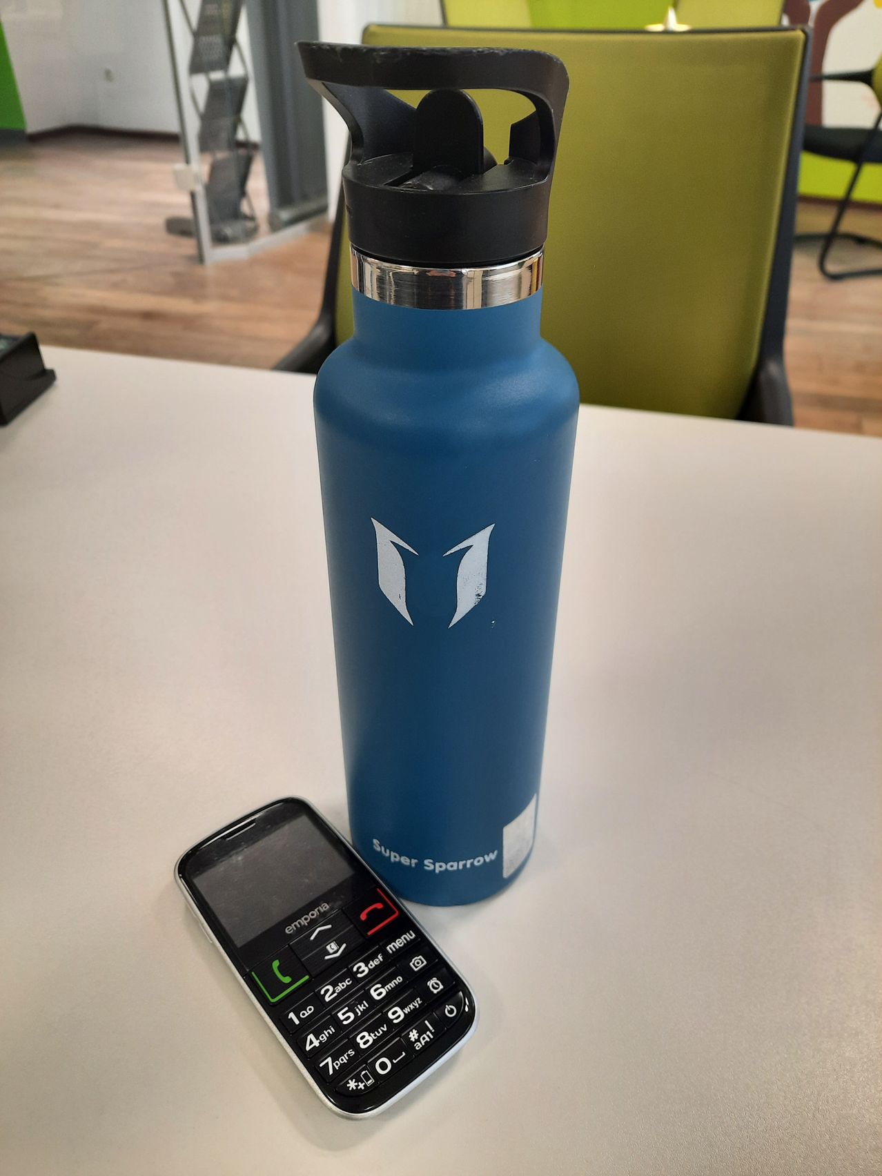 Eine blaue Super Sparrow Flasche steht auf einem weißen Tisch. Daneben befindet sich ein schwarzes Telefon, auf dessen Display das Menü zu sehen ist.
