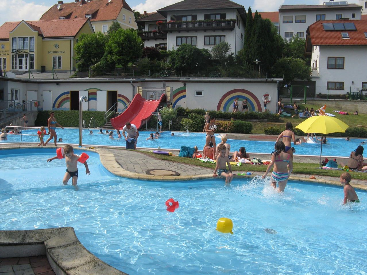 Ein großes Außenbecken mit Regenbogenwand, Rutsche und Schwimmenden. Mehrere Kinder spielen und planschen im Wasser. Im Hintergrund befinden sich Gebäude und Bäume.