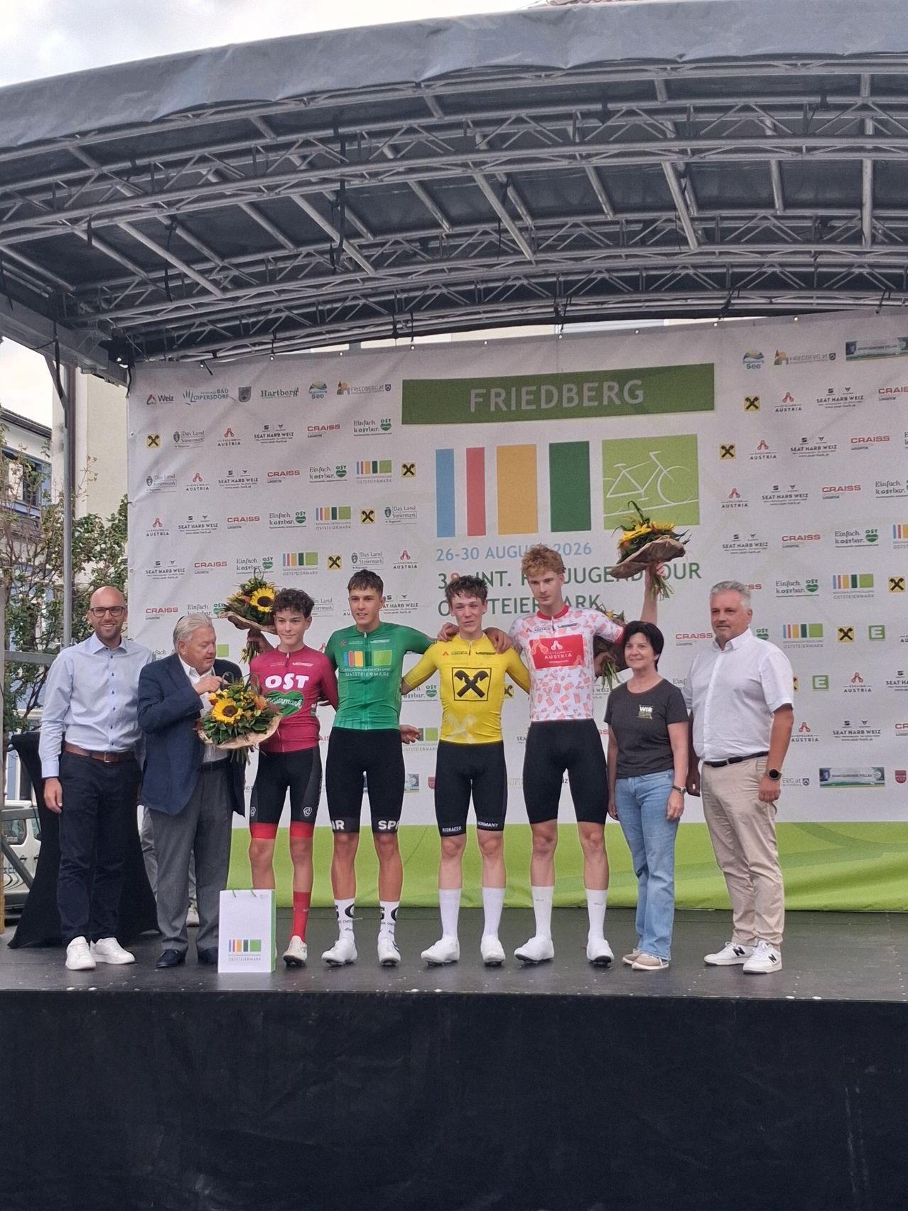 Five cyclists stand on a podium, wearing cycling jerseys and holding bouquets of flowers. Behind them, a banner displays the event name 'Friedberg'. Two men and three women stand beside them, all smiling.