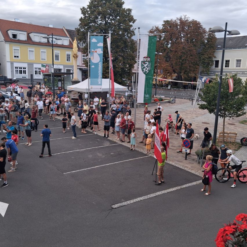A crowd of people gather in an outdoor area, possibly for an event. Some are standing, while others are walking. There are flags and banners on poles, and a white tent is set up. Buildings, trees, and streetlights are in the background.