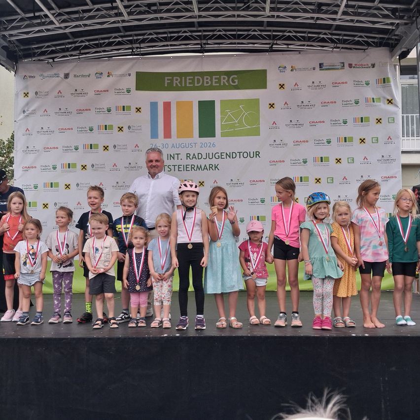 A group of children and an adult man are standing on a stage, wearing medals and smiling for a photo. Behind them is a banner with the text 'Friedberg' and 'Int. Radjugendtour Steiermark'.